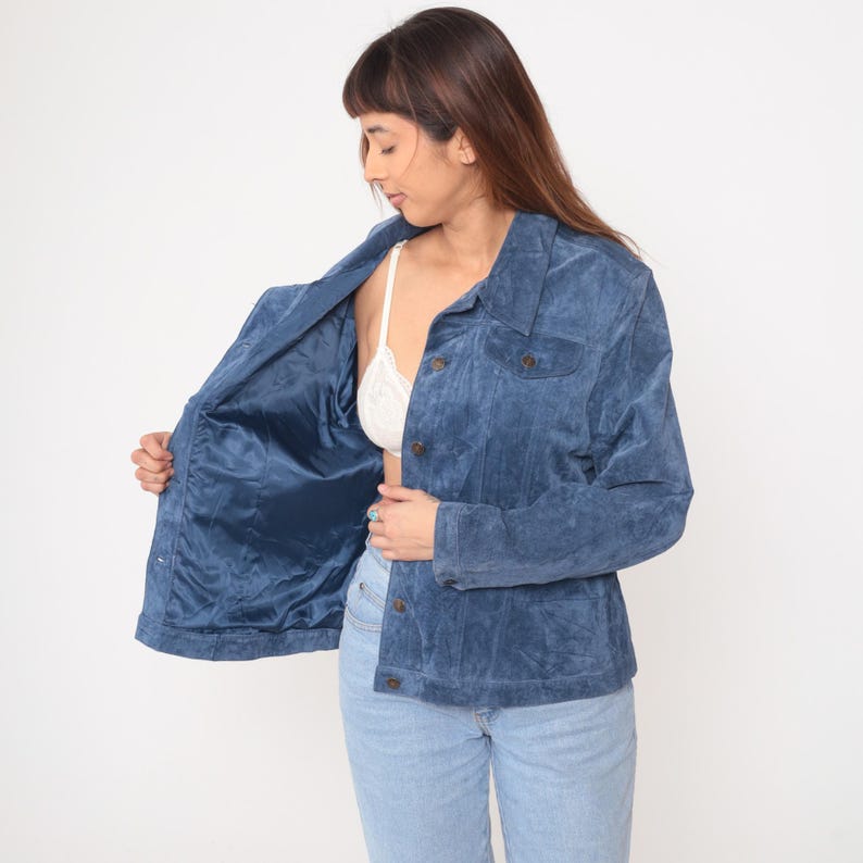 May include: A blue suede jacket with a button-up front and two chest pockets. The jacket is open, revealing a blue satin lining. The person is wearing light blue jeans and a white bra.