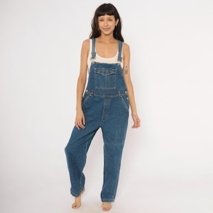 Könnte beinhalten: Blaue Jeans-Latzhose mit verstellbaren Trägern, einer Brusttasche und Seitentaschen. Die Latzhose hat eine lockere Passform und ist aus mittelblauem Denim gefertigt. Das Model trägt ein weißes Tanktop darunter. Ein klassisches, lässiges Kleidungsstück.