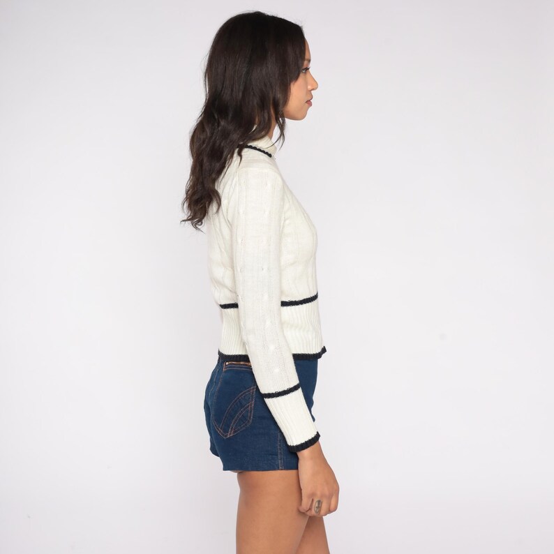 May include: Cream-colored cable knit sweater with black trim on the collar, cuffs, and waist. The long-sleeved sweater is paired with dark blue denim shorts. The outfit is shown from a side view.