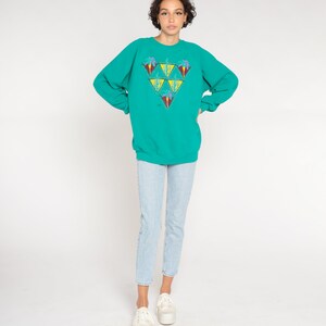 May include: Teal sweatshirt with a geometric design of triangles, each containing a palm tree and other colorful elements. The sweatshirt is paired with light blue jeans and white platform sneakers.