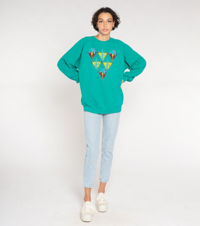 May include: Teal sweatshirt with a geometric design of triangles, each containing a palm tree and other colorful elements. The sweatshirt is paired with light blue jeans and white platform sneakers.