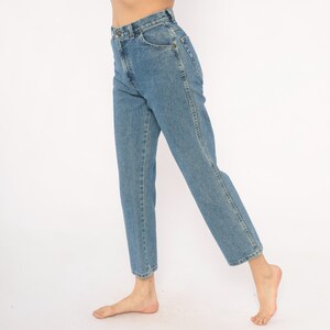 May include: Light blue denim jeans with a classic straight leg fit. The jeans have a button and zipper closure, front and back pockets, and a faded wash. The model is wearing the jeans with bare feet.
