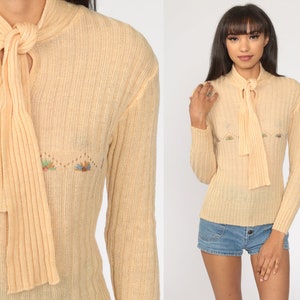 May include: A pale yellow knitted sweater with a long tie at the neck and embroidered floral details. The sweater has long sleeves and a ribbed texture.