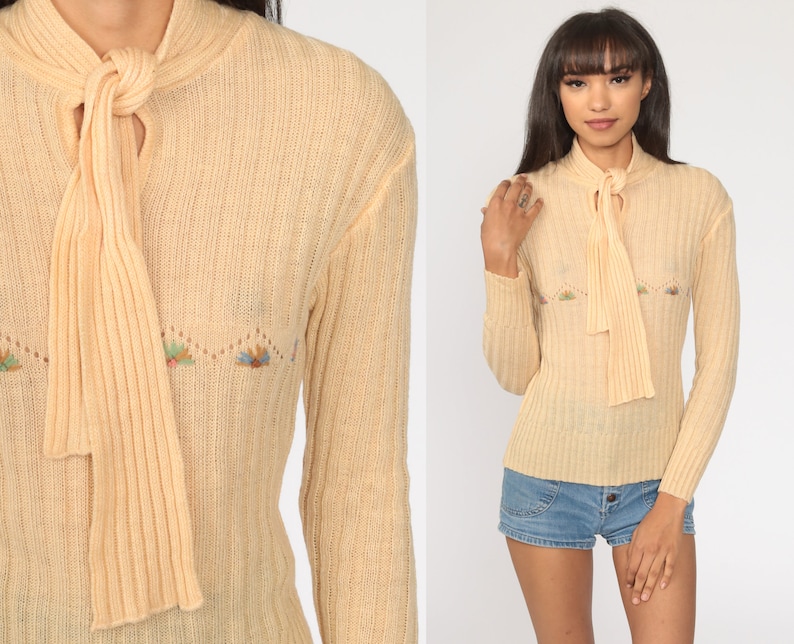 May include: A pale yellow knitted sweater with a long tie at the neck and embroidered floral details. The sweater has long sleeves and a ribbed texture.