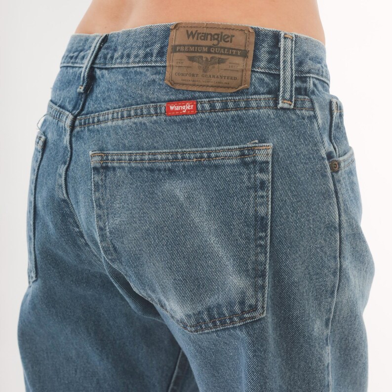 May include: Light blue denim jeans with a classic five-pocket design. The back features a Wrangler brand patch and a red Wrangler tag on the back pocket. The jeans have a straight leg cut.