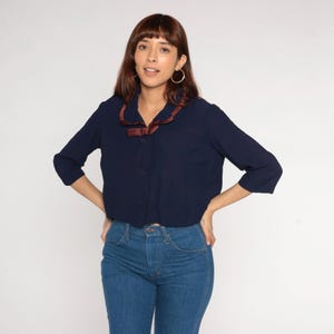May include: A navy blue blouse with a brown satin collar and a matching bow. The blouse has three-quarter sleeves and is buttoned down the front. The model is wearing blue jeans.
