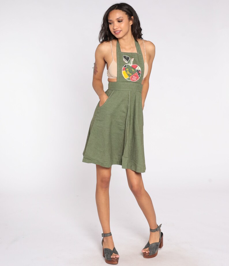 May include: Olive green apron dress with an apple applique. The dress features a halter neckline, pockets, and a flared skirt. The apple applique is made of various colorful fabric patches. The model is wearing gray sandals with wooden heels.