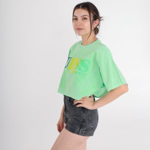 May include: A light green cropped t-shirt with a faded rainbow coloured "GUESS" logo. The shirt has short sleeves and a relaxed fit.