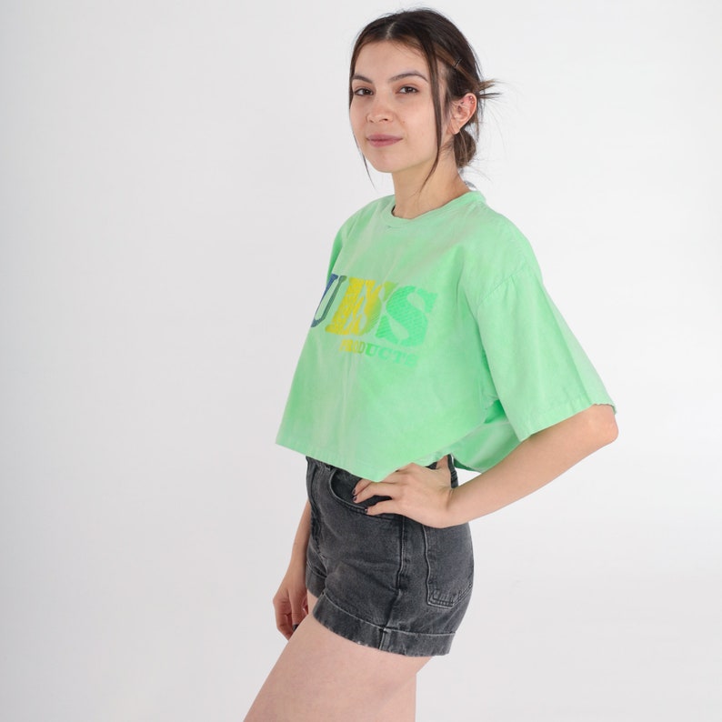 May include: A light green cropped t-shirt with a faded rainbow coloured "GUESS" logo. The shirt has short sleeves and a relaxed fit.