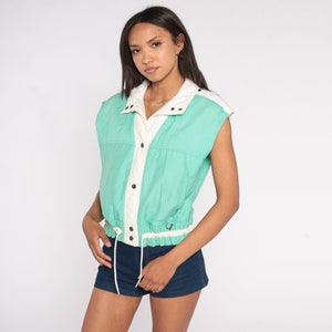 K&ouml;nnte beinhalten: Eine &auml;rmellose Weste in Mintgr&uuml;n und Off-White. Die Weste hat einen Kordelzug in der Taille, Druckkn&ouml;pfe und einen Kragen. Das Kleidungsst&uuml;ck ist aus leichtem Material gefertigt und eignet sich perfekt zum &Uuml;berziehen. Die Weste ist im Vintage-Stil.