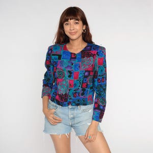 Könnte beinhalten: Eine langärmlige Bluse mit einem lebendigen Patchwork-Design. Die Bluse hat eine Mischung aus blauen, rosa und lila Quadraten mit abstrakten Mustern. Sie hat einen runden Ausschnitt und wird mit Jeansshorts getragen.