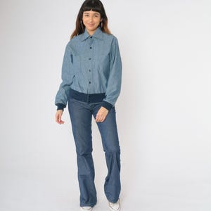 May include: A blue denim shirt with a button-up front and a navy blue ribbed collar and cuffs. The shirt is worn with blue jeans.