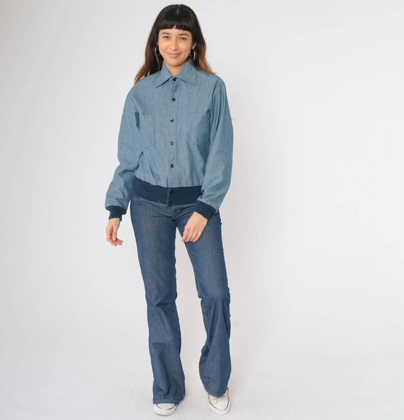 May include: A blue denim shirt with a button-up front and a navy blue ribbed collar and cuffs. The shirt is worn with blue jeans.