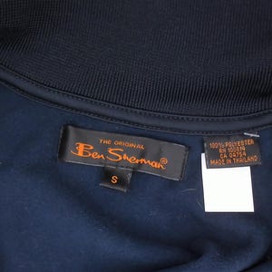 May include: A dark navy blue garment featuring a ribbed collar and a black interior label with the orange text The Original Ben Sherman and a size S tag. A secondary black tag displays 100% Polyester, RN 100819, CA 04754, and Made in Thailand.