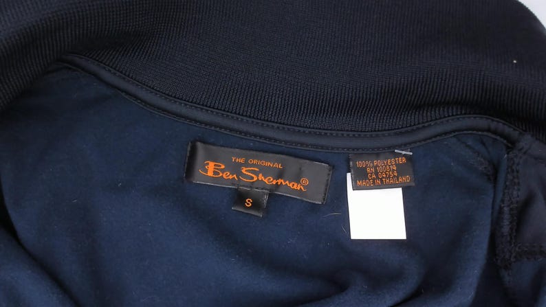 May include: A dark navy blue garment featuring a ribbed collar and a black interior label with the orange text The Original Ben Sherman and a size S tag. A secondary black tag displays 100% Polyester, RN 100819, CA 04754, and Made in Thailand.