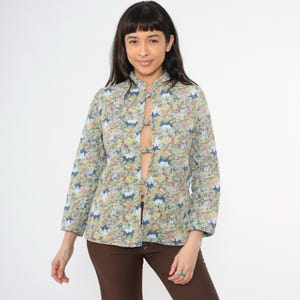 May include: A light blue floral print button-up shirt with a mandarin collar and a long sleeve. The shirt has a unique design with a center opening and decorative buttons.