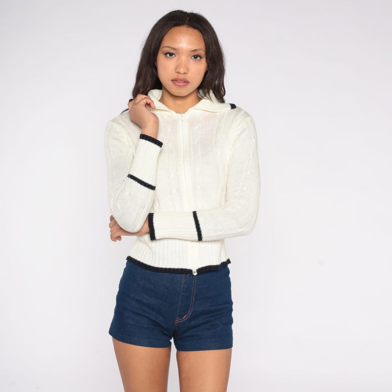 May include: Cream-colored zip-up sweater with navy blue trim on the collar, cuffs, and hem. The sweater has a cable knit design and is paired with dark blue denim shorts. The outfit is a vintage style.