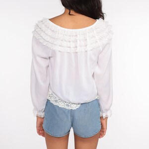 May include: White long-sleeved blouse with a ruffled lace neckline and lace trim at the cuffs and hem. The blouse is worn with denim shorts.