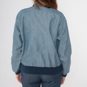 May include: A light blue denim jacket with a navy blue ribbed knit collar and cuffs. The jacket has a button closure and long sleeves.