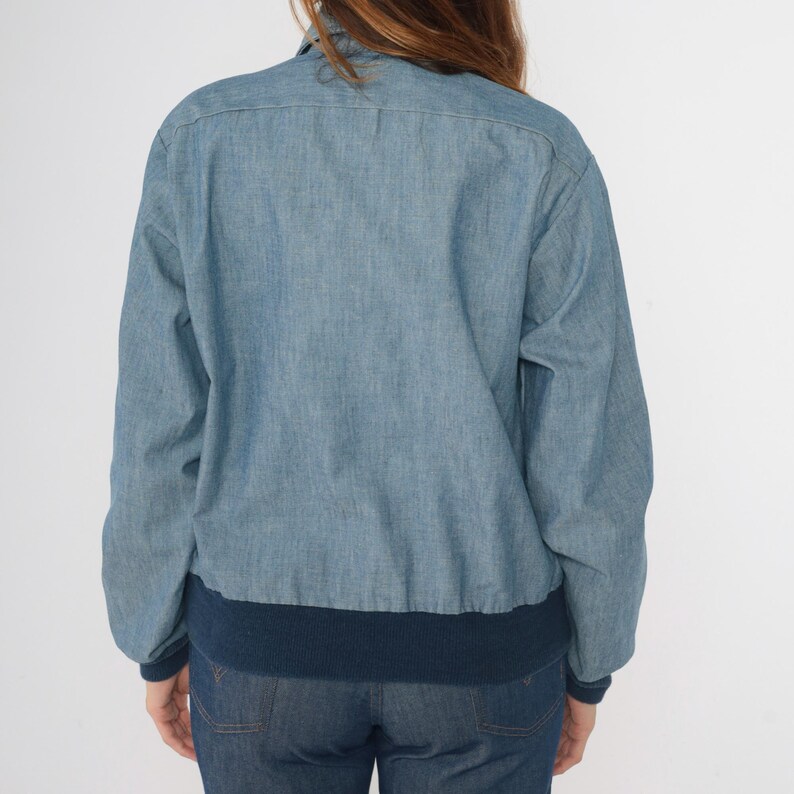May include: A light blue denim jacket with a navy blue ribbed knit collar and cuffs. The jacket has a button closure and long sleeves.