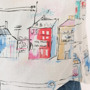 May include: A white shirt featuring a colorful hand-drawn style print of a town street. The design includes buildings labeled Fox Tavern Free House, The Ship, and Swan, with black ink outlines and accents in red, blue, yellow, and pink watercolor tones.