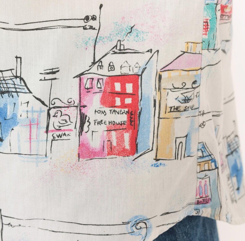 May include: A white shirt featuring a colorful hand-drawn style print of a town street. The design includes buildings labeled Fox Tavern Free House, The Ship, and Swan, with black ink outlines and accents in red, blue, yellow, and pink watercolor tones.