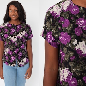 May include: A short-sleeved button-up shirt with a black background and a floral pattern of purple and white roses. The shirt has a round collar and is made of a lightweight fabric.