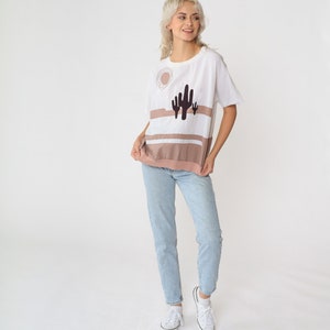 May include: A white t-shirt with a brown and white striped design and a cactus graphic. The shirt has a relaxed fit and short sleeves.