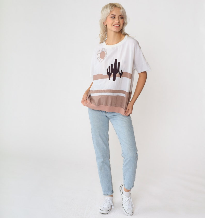 May include: A white t-shirt with a brown and white striped design and a cactus graphic. The shirt has a relaxed fit and short sleeves.