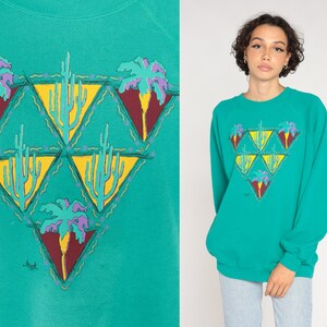 May include: Teal sweatshirt with a desert-themed graphic. The design features yellow triangles with cacti and palm trees in purple, yellow, and maroon. The sweatshirt has long sleeves and a crew neck.