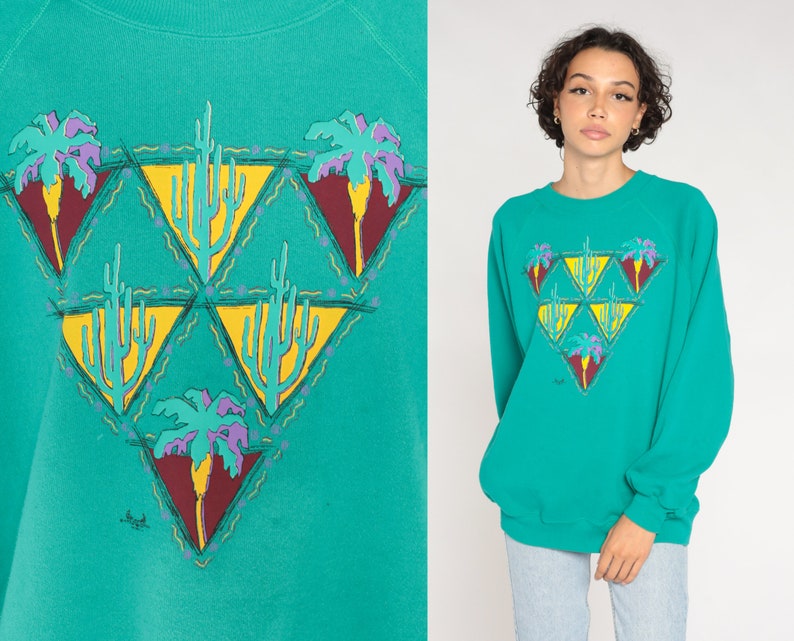 May include: Teal sweatshirt with a desert-themed graphic. The design features yellow triangles with cacti and palm trees in purple, yellow, and maroon. The sweatshirt has long sleeves and a crew neck.