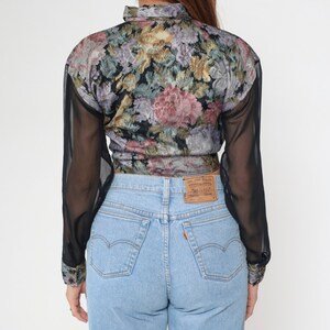 May include: A vintage floral print blouse with sheer black sleeves and light blue denim jeans. The blouse features a colorful floral pattern with pink, purple, and yellow flowers on a black background. The jeans have a classic five-pocket design.