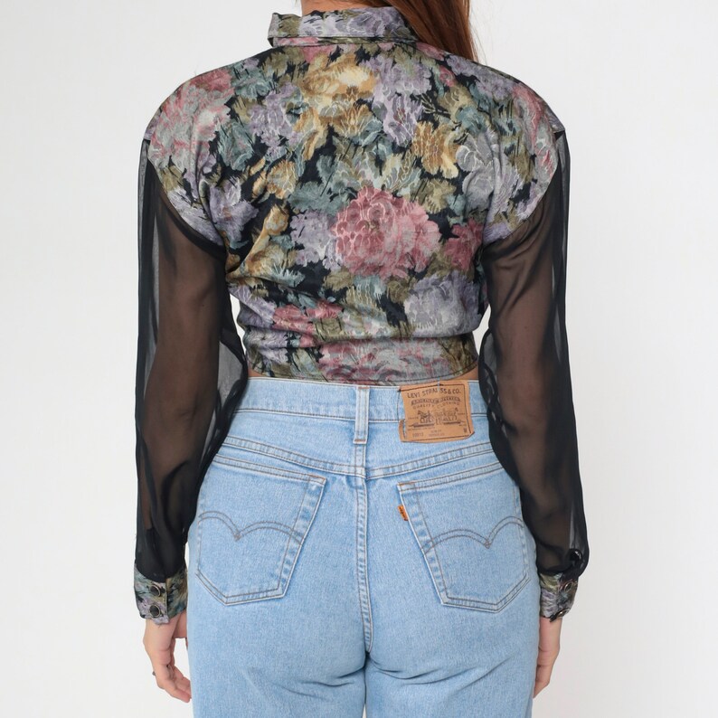 May include: A vintage floral print blouse with sheer black sleeves and light blue denim jeans. The blouse features a colorful floral pattern with pink, purple, and yellow flowers on a black background. The jeans have a classic five-pocket design.