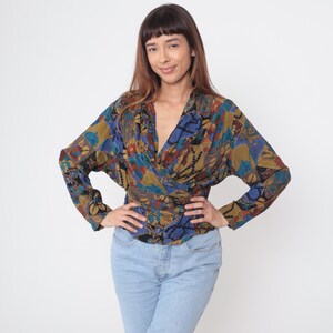 May include: A patterned wrap blouse in shades of blue, brown, and gold. The long-sleeved top has a V-neck and a wrap design. The blouse is paired with light blue jeans.