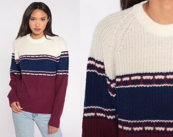 May include: A cream-colored sweater with a crew neck and long sleeves. The sweater features a navy blue band with white and burgundy stripes across the chest and a burgundy lower section. The sweater is made of a thick knit material.