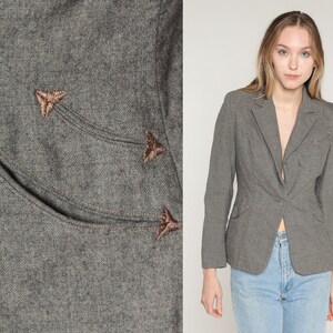 May include: A gray blazer with a single-button closure and two front pockets. The blazer features decorative stitching and embroidered arrow-shaped accents in a contrasting color. The model is wearing blue jeans.