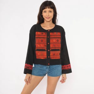 May include: A black long-sleeve jacket featuring a central panel and sleeve cuffs with a red and black woven geometric pattern. The jacket has a round neckline, a front button closure with red buttons, and two horizontal pockets on the front.