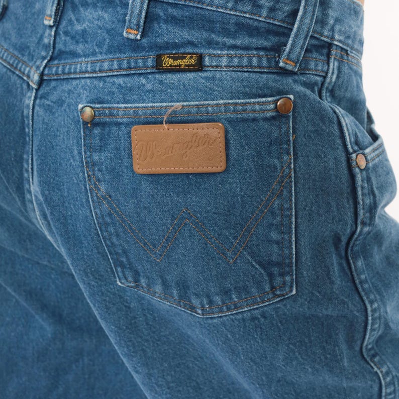 May include: Close-up of blue denim jeans with a Wrangler label on the back pocket. The jeans have a classic five-pocket design with copper-colored rivets and stitching. The Wrangler label is a brown leather rectangle.