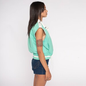 K&ouml;nnte beinhalten: Eine &auml;rmellose Vintage-Weste in T&uuml;rkis und Wei&szlig; mit Kordelzug in der Taille. Die Weste hat einen wei&szlig;en Kragen und einen wei&szlig;en Streifen am unteren Saum. Das Model tr&auml;gt Jeansshorts.