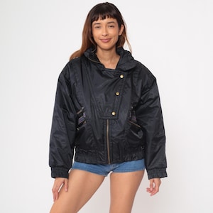 May include: Black nylon jacket with a high collar, gold-colored buttons, and a zipper closure. The jacket has a slightly oversized fit and features decorative patterned fabric on the pockets. The jacket is a vintage style.