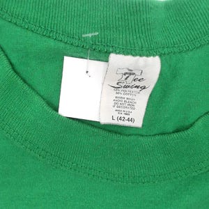 K&ouml;nnte beinhalten: Nahaufnahme eines gr&uuml;nen T-Shirts mit einem wei&szlig;en Etikett. Auf dem Etikett steht "7 Tee Swing" und enth&auml;lt Waschanweisungen und die Gr&ouml;&szlig;e L (42-44). Das Shirt besteht aus 50 % Polyester und 50 % Baumwolle.
