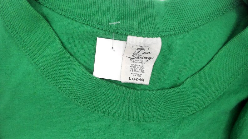 K&ouml;nnte beinhalten: Nahaufnahme eines gr&uuml;nen T-Shirts mit einem wei&szlig;en Etikett. Auf dem Etikett steht "7 Tee Swing" und enth&auml;lt Waschanweisungen und die Gr&ouml;&szlig;e L (42-44). Das Shirt besteht aus 50 % Polyester und 50 % Baumwolle.