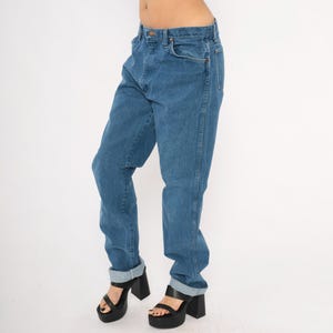 May include: Blue denim jeans with a classic five-pocket design. The jeans have a straight leg fit and are cuffed at the ankles. The model is wearing black platform heels. The jeans are a medium wash.