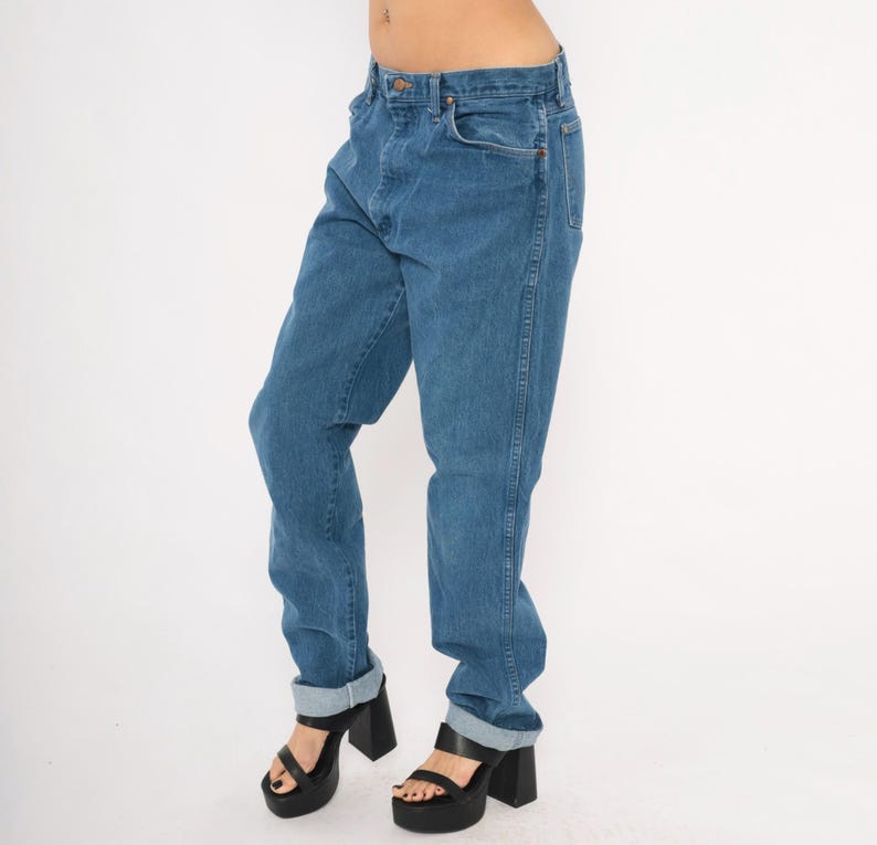 May include: Blue denim jeans with a classic five-pocket design. The jeans have a straight leg fit and are cuffed at the ankles. The model is wearing black platform heels. The jeans are a medium wash.