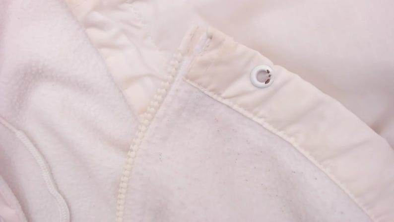 Puede incluir: Primer plano de una chaqueta blanca con forro polar. La chaqueta tiene cremallera y capucha. La tela es suave y texturizada. Se ve un ojal met&aacute;lico en la capucha.