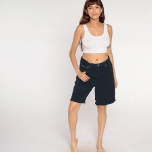 Puede incluir: Una camiseta sin mangas blanca y unos shorts vaqueros azul oscuro. Los shorts tienen un dobladillo sin rematar y cierre de botón y cremallera. La modelo lleva pendientes de aro dorados.