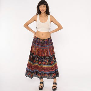 May include: A long, tiered skirt with a vibrant floral pattern in red, blue, yellow, and black. The skirt has a drawstring waist and is paired with a white tank top. The model is wearing black platform sandals.