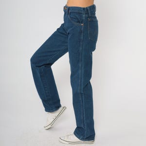 May include: Dark blue denim jeans with a classic straight-leg fit. The jeans feature a traditional five-pocket design, a button and zipper closure, and copper-colored rivets. The model is wearing white sneakers.