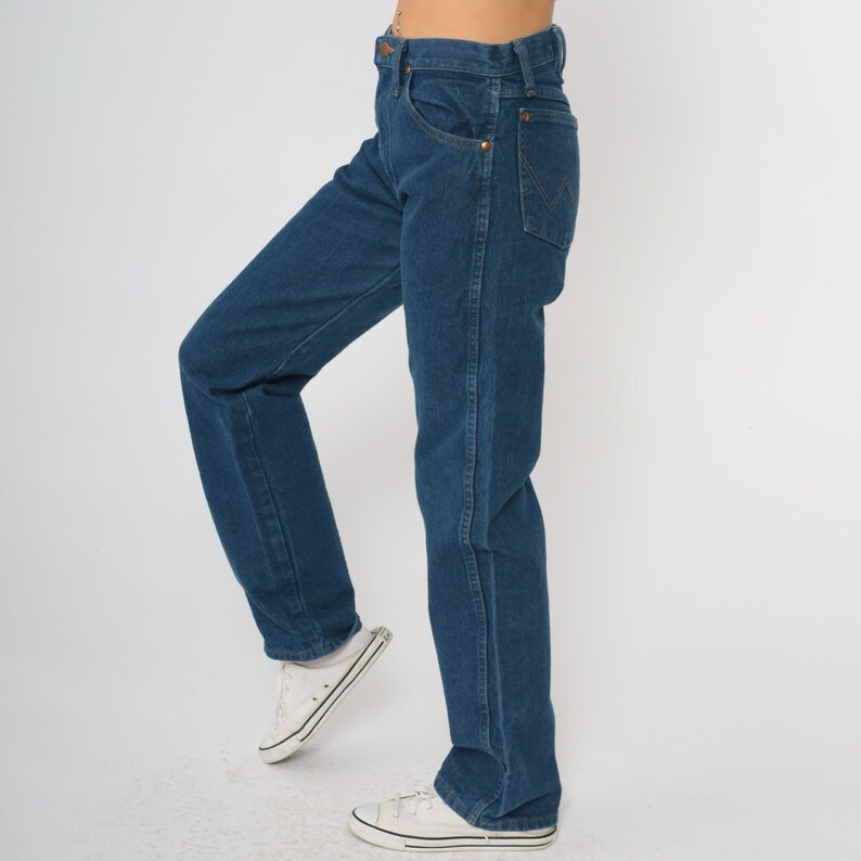 May include: Dark blue denim jeans with a classic straight-leg fit. The jeans feature a traditional five-pocket design, a button and zipper closure, and copper-colored rivets. The model is wearing white sneakers.