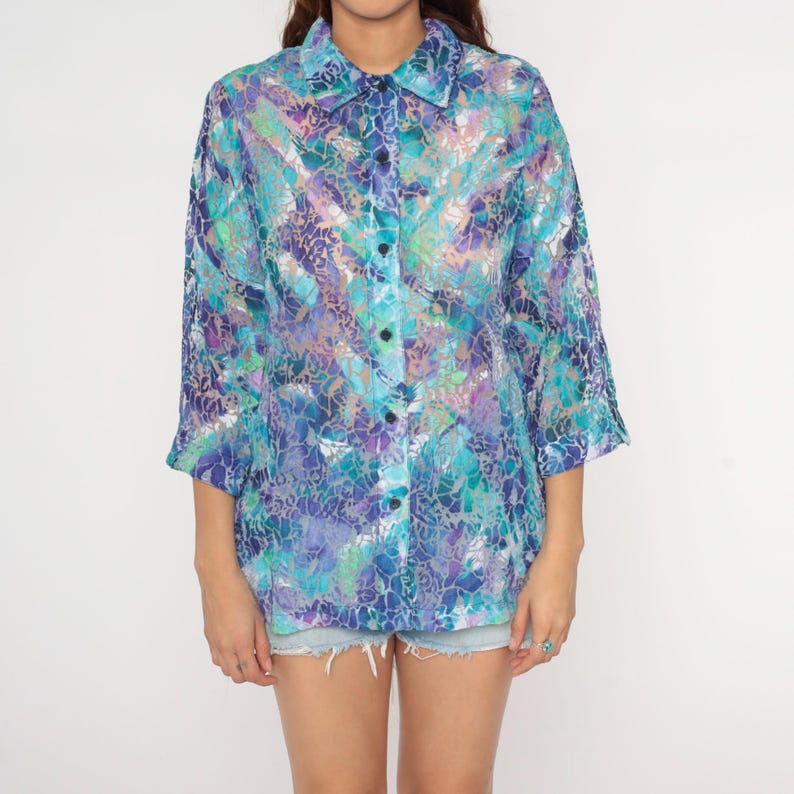 Op de afbeelding: Een transparante blouse met knoopsluiting en een abstract patroon in blauw, paars en groen. De blouse heeft een kraag en driekwart mouwen. De lichte, semi-transparante stof zorgt voor een stijlvolle uitstraling. Perfect voor diverse gelegenheden.